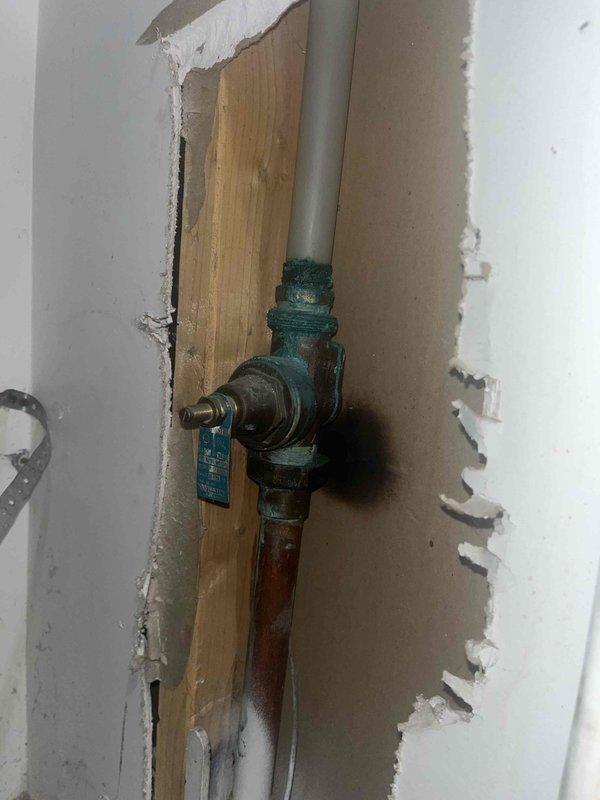 Responded to no hot water complaint on 6-year-old tanked water heater in basement. Upon inspection, observed water heater with exposed shut-off valve assembly and heating element through viewing window showing active ignition. Documented existing plumbing configuration including supply lines and main shut-off valve mounted to access panel. Inspected heating element operation and verified gas supply connections through access port, noting proper ignition sequence with visible flame present.