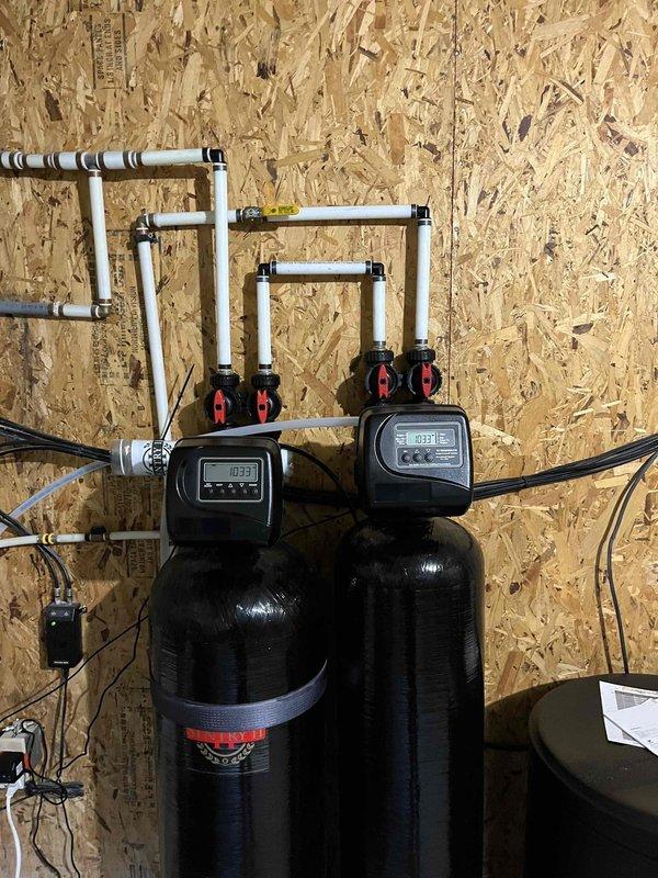 Installed a complete whole-home water treatment system consisting of a Pacific Water Sentry 2 salt-free water conditioner paired with a complementary filtration unit. Mounted two black cylindrical treatment tanks side-by-side against utility room wall with digital control heads featuring LCD displays and manual valve controls. Configured white PVC manifold piping system across the top of both units with multiple connection points, vertical drops, and elbow fittings to integrate with existing plumbing infrastructure. Connected electrical components including power adapters and control wiring to wall outlets for system operation. Integrated the installation with existing copper supply lines, gray PEX tubing, and HVAC components in the mechanical room. The Sentry 2 system provides maintenance-free water conditioning capable of treating up to one million gallons over its operational lifespan, removing hydrogen sulfide, chlorine, fluoride, pesticides, sediments, and heavy metals beyond stan