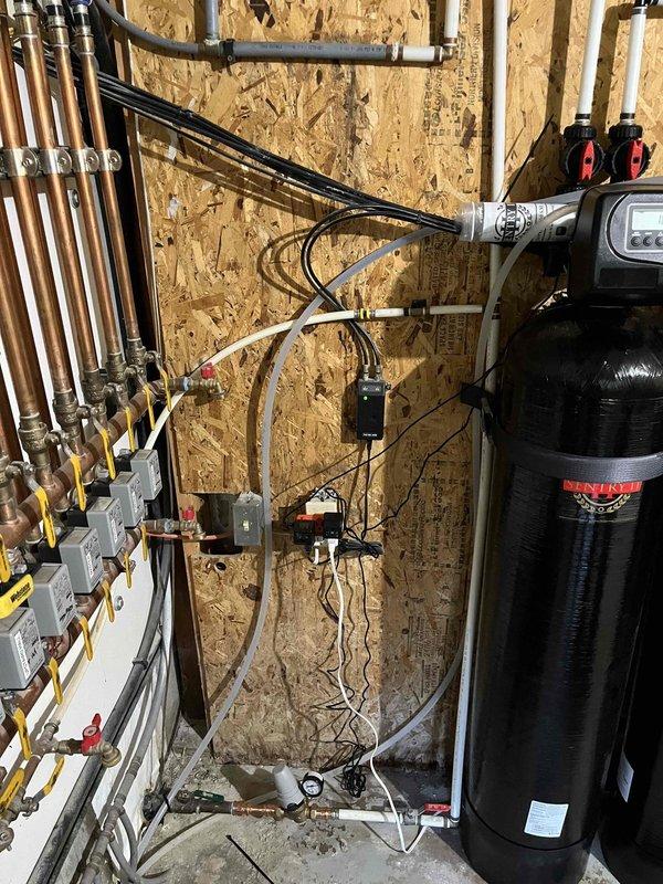 Installed a complete whole-home water treatment system consisting of a Pacific Water Sentry 2 salt-free water conditioner paired with a complementary filtration unit. Mounted two black cylindrical treatment tanks side-by-side against utility room wall with digital control heads featuring LCD displays and manual valve controls. Configured white PVC manifold piping system across the top of both units with multiple connection points, vertical drops, and elbow fittings to integrate with existing plumbing infrastructure. Connected electrical components including power adapters and control wiring to wall outlets for system operation. Integrated the installation with existing copper supply lines, gray PEX tubing, and HVAC components in the mechanical room. The Sentry 2 system provides maintenance-free water conditioning capable of treating up to one million gallons over its operational lifespan, removing hydrogen sulfide, chlorine, fluoride, pesticides, sediments, and heavy metals beyond stan