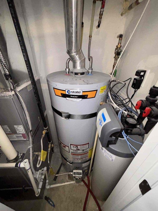 Performed water heater flush service on a 6-year-old State brand tank-type water heater located in the utility room accessible through the garage. Inspected all plumbing connections, including copper supply lines, PVC drainage piping, and flue venting system, confirming proper installation and operation of water heater alongside adjacent water treatment system with functioning valve assemblies.