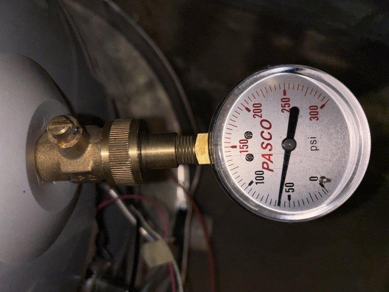 Inspected the plumbing supplying the tanked water heater in response to a reported pipe leak and found corrosion and mineral buildup (verdigris and scale) at the brass fittings on both sides of the pressure-reducing valve, indicating a leak point at the threaded joint and clamped connection. Verified system pressure with an analog gauge at approximately 50 psi, within normal operating range. Documented the condition of the fittings and recommended repair or replacement of the corroded connections to prevent further leakage.