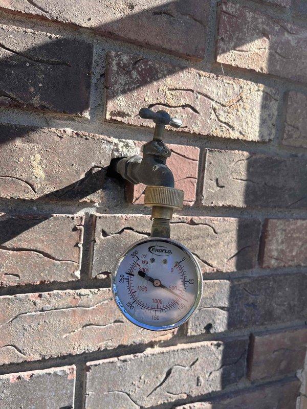 Inspected the kitchen sink area in response to a reported faucet leak with water damage to the adjacent wall, and connected a pressure gauge to the exterior hose bib to test static water pressure. The gauge read approximately 250 psi, far above the normal residential range of 40–80 psi, indicating excessive supply pressure that likely contributed to the leak and puts fixtures and piping at risk of further failure. Documented the findings with photos; a pressure-reducing valve and repair of the leaking fixture/piping are recommended, and the leak is causing damage (Damage tag applies).