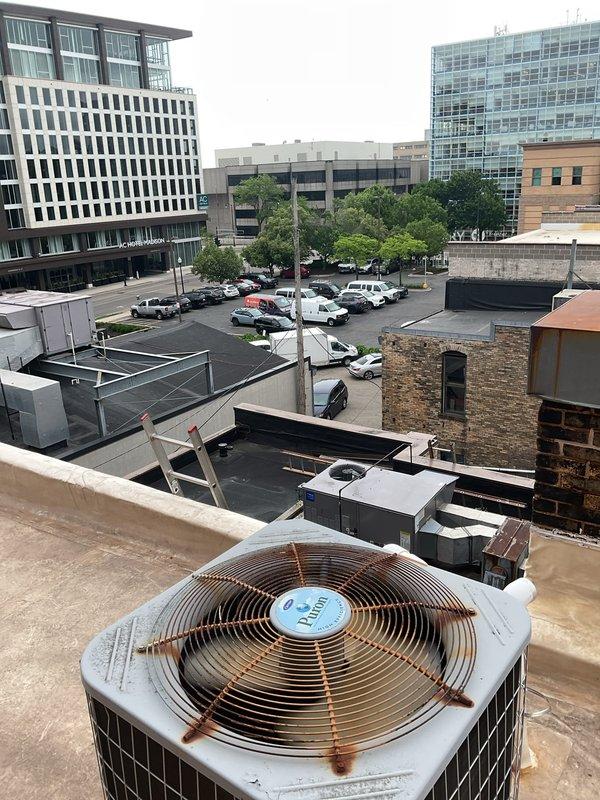 Responded to service call for leaking rooftop AC unit located on commercial building fifth floor, accessed through YWCA facility with tenant code assistance. Upon inspection, found white HVAC condensing unit with weathered components and visible rust on fan grille, connected to black refrigerant lines and gray electrical disconnect. Identified condensate drainage issue with white PVC piping system showing discoloration at roof membrane interface. Documented rooftop equipment configuration including multiple commercial air handling units and assessed condition of refrigerant line connections and condensate management system to address reported water leakage.