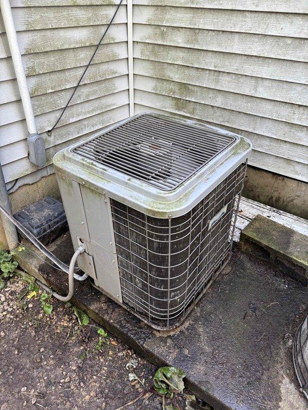 Responded to no cooling complaint on aging residential air conditioning system. Upon arrival, found outdoor condenser unit showing significant signs of age and weathering, with visible algae and mold growth on top grille and casing. Unit located in corner installation against house exterior with white vinyl siding also exhibiting organic growth and environmental deterioration.

Performed visual inspection of equipment exterior and surrounding installation area. Documented overall condition of condensing unit including weathered metal casing, discolored protective grille, and compromised appearance of refrigerant line insulation. Noted electrical disconnect box and conduit routing. Equipment showed signs of extended service life with minimal recent maintenance history based on visible accumulation of organic growth and general weathering patterns.

Captured photographic documentation of unit condition, installation configuration, and surrounding environmental factors affecting equipment