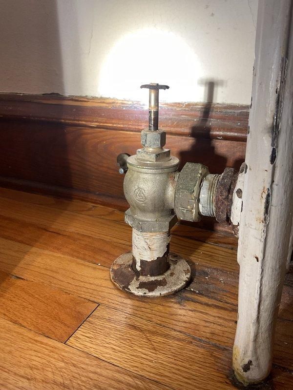 Responded to a complaint of excessive steam and uncontrolled heat output from the bedroom radiator nearest the front entrance, causing moisture on the floor. Inspected the radiator steam valve and found the valve, union fitting, and floor flange heavily corroded, and the radiator air vent mounted to the frame showing rust staining. Serviced the radiator valve and venting components to restore proper steam control and prevent further moisture release.