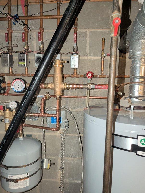 Completed installation of a residential hydronic heating system featuring copper pipe network with multiple zone valves and control modules. System includes a green circulator pump, pressure regulation assembly with gauges, and properly connected expansion tank. All components were installed to code, pressure tested, and verified for optimal operation.