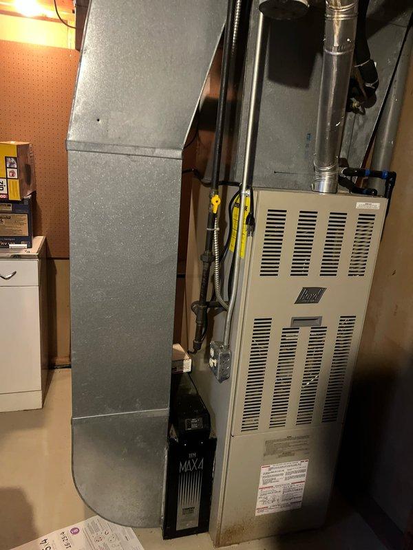 Inspected B-vent pipe system in basement/utility room and identified active leakage. Accessed installation via ladder through garage entry point. System consists of white furnace unit connected to vertical metal ductwork with proper ventilation components. Recommended sealing or replacement of compromised B-vent sections to prevent further leakage and ensure safe operation.