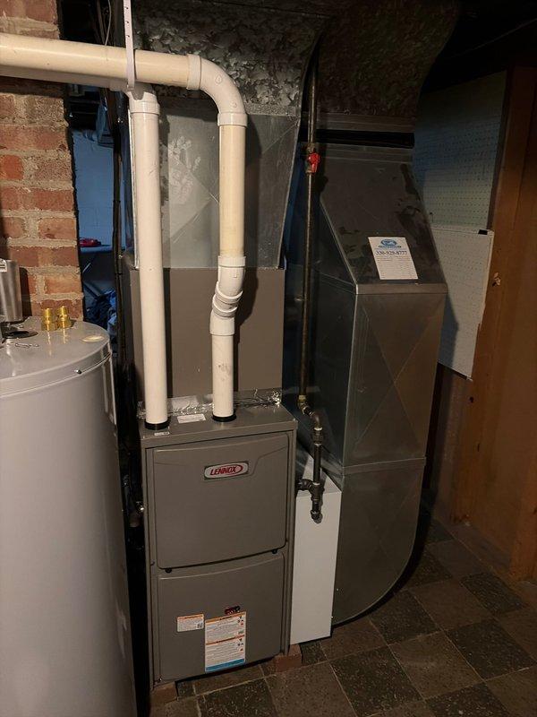 Completed Lennox furnace retrofit installation in basement mechanical room, including proper PVC venting through exterior wall. Installed unit features standard manufacturer specifications with appropriate connections to existing HVAC system and water heater. Exterior penetration properly sealed despite evidence of pre-existing moisture issues on siding.