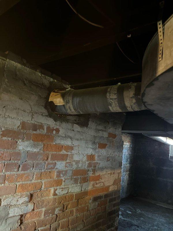 Inspected deteriorated ductwork in basement area showing significant rust staining and compromised insulation wrap. Observed moisture damage affecting the cylindrical duct system mounted along brick/stone foundation wall, with visible yellowing and deterioration of insulation material requiring professional remediation.
