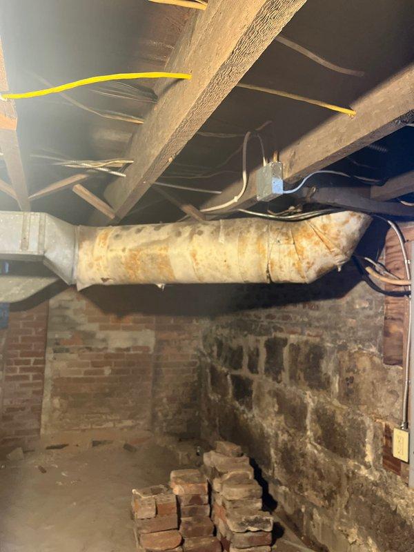 Inspected deteriorated ductwork in basement area showing significant rust staining and compromised insulation wrap. Observed moisture damage affecting the cylindrical duct system mounted along brick/stone foundation wall, with visible yellowing and deterioration of insulation material requiring professional remediation.