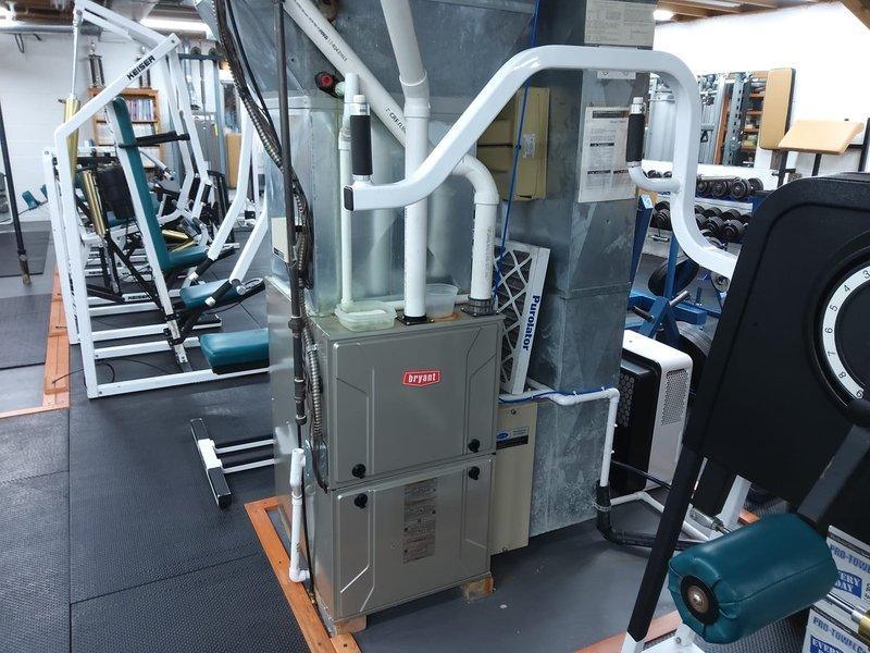 Conducted an AC retrofit assessment in a basement where a Bryant furnace system is currently installed on a wooden platform. Documented the existing HVAC setup including the furnace unit, PVC exhaust pipes, and surrounding metal ductwork components to prepare for air conditioning integration with the existing system.