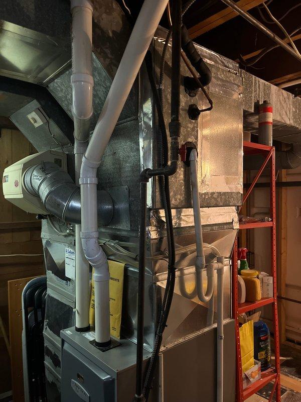 Completed full AC system retrofit including outdoor condenser unit and indoor air handler replacement. Installed new condenser unit on exterior pad adjacent to light green siding with proper clearances, mounted electrical disconnect box on exterior wall, and ran conduit connections to unit. Interior installation included new air handler in basement utility area with complete PVC condensate drain piping in Y-configuration, insulated supply plenum, flexible return ductwork connections, and refrigerant line set routing between indoor and outdoor units. Verified all electrical connections, tested system startup and operation, and confirmed proper drainage flow.