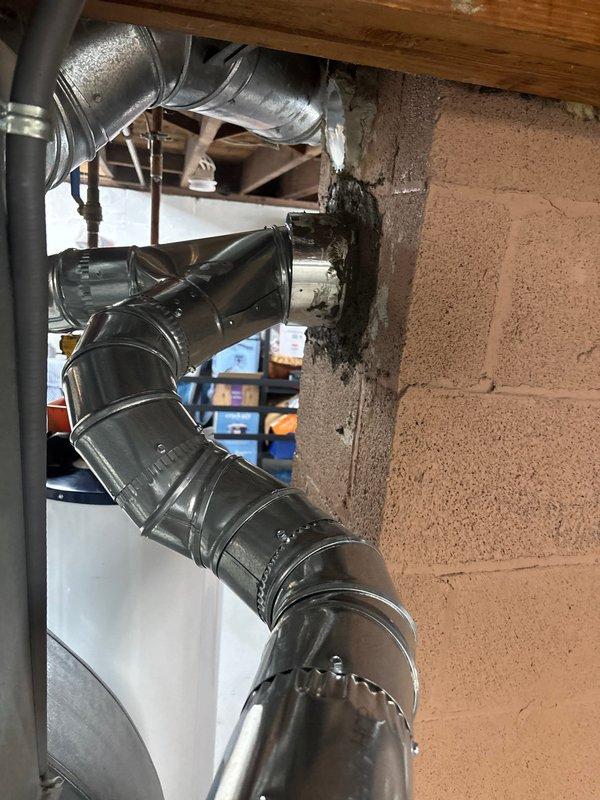 Installed flexible metallic liner material through existing masonry wall penetration to connect HVAC ductwork between utility area and adjacent space. Secured all flexible duct connections with metal clamps and banding to ensure proper airflow and system integrity. Documented completed installation with photographs showing proper routing and connection of black cylindrical duct sections to galvanized ductwork through basement wall opening.