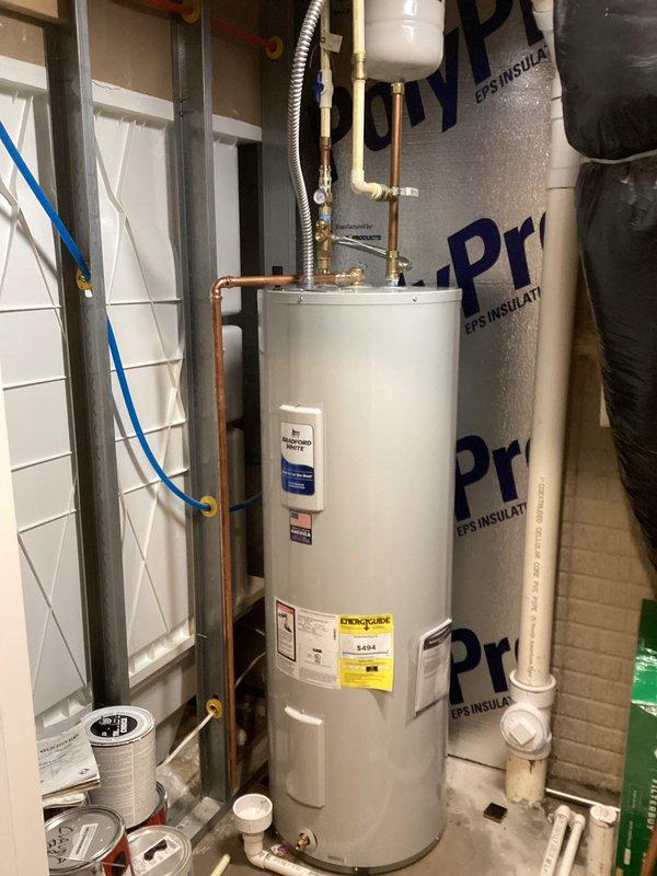 Installed new white cylindrical tanked water heater in residential utility room. Connected copper supply lines with brass fittings and flexible metal connector at top of unit, installed white expansion tank above water heater, and completed all necessary plumbing connections including shut-off valves at base of unit. Unit properly positioned with reflective insulation backing and all manufacturer labels visible for future reference.