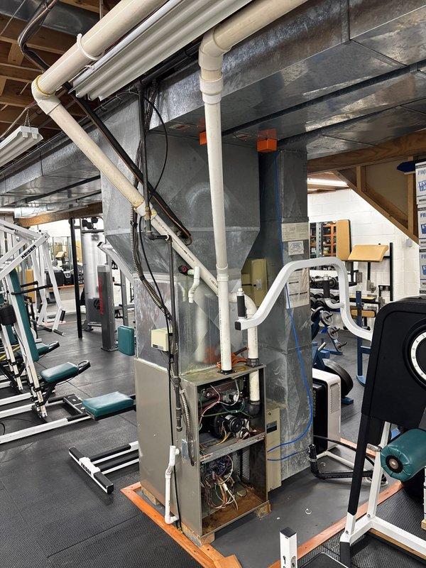 Completed full HVAC system installation in commercial gym facility, including main air handling unit with integrated ductwork, zoning controls, and humidifier. The system features exposed ceiling-mounted galvanized metal ductwork with funnel-style plenum configuration, PVC condensate drainage lines, electrical conduit routing, and copper refrigerant lines, all installed to accommodate the facility's open-ceiling design while maintaining functionality around existing exercise equipment.