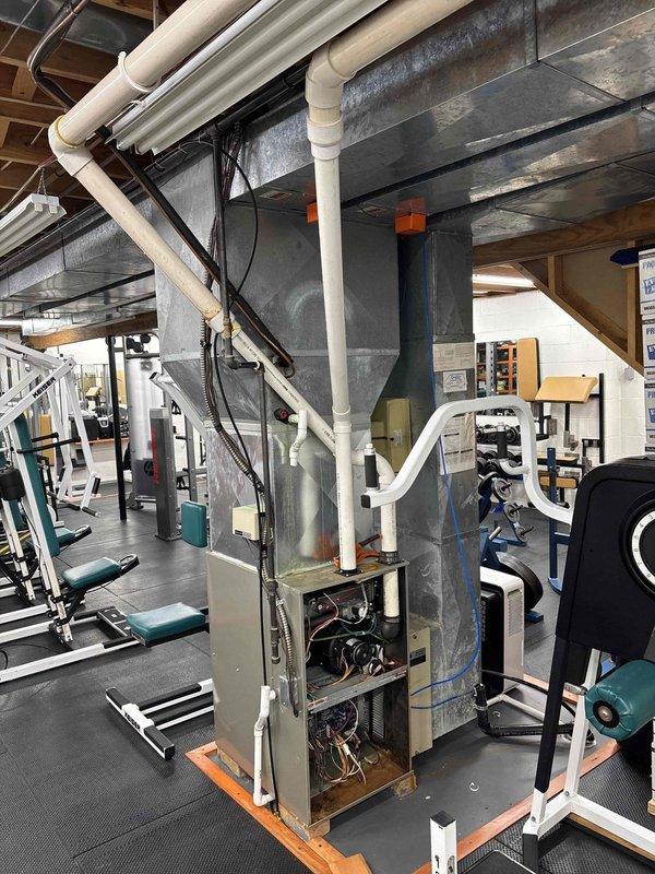 Completed full HVAC system installation in commercial gym facility, including main air handling unit with integrated ductwork, zoning controls, and humidifier. The system features exposed ceiling-mounted galvanized metal ductwork with funnel-style plenum configuration, PVC condensate drainage lines, electrical conduit routing, and copper refrigerant lines, all installed to accommodate the facility's open-ceiling design while maintaining functionality around existing exercise equipment.