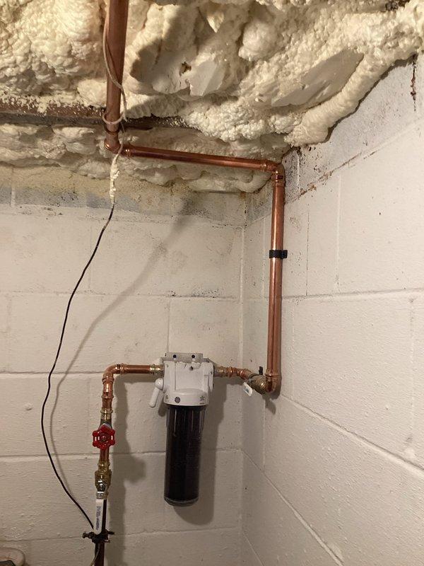 Installed customer-supplied whole house water filtration system with complete rework of existing copper water lines. The installation included mounting a black cylindrical filter housing to the basement wall, integrating a red-handled shutoff valve and pressure regulation device, and reconfiguring copper piping with brass fittings to accommodate the new filtration system while maintaining proper water flow throughout the residence.