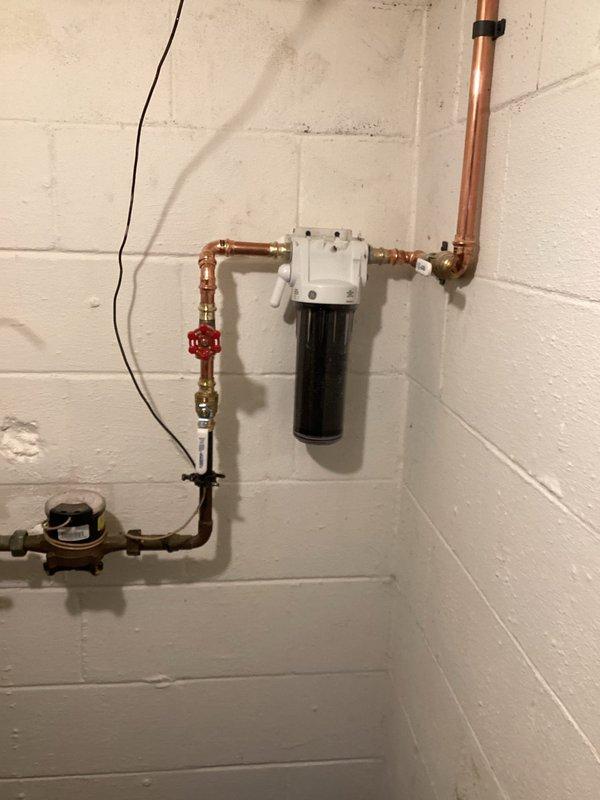 Installed customer-supplied whole house water filtration system with complete rework of existing copper water lines. The installation included mounting a black cylindrical filter housing to the basement wall, integrating a red-handled shutoff valve and pressure regulation device, and reconfiguring copper piping with brass fittings to accommodate the new filtration system while maintaining proper water flow throughout the residence.