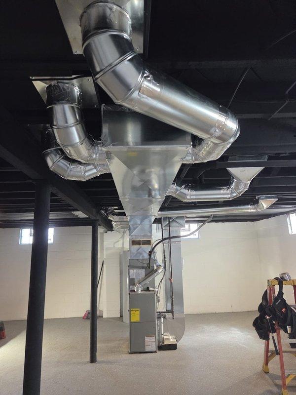 Completed new construction HVAC ductwork installation in basement mechanical room, including full trunk line and branch duct system fabricated from galvanized metal with proper hangers and support strapping secured to ceiling joists. Installed and positioned gray furnace/air handler unit on concrete floor with all necessary connections, ensuring proper clearances and access for future maintenance per manufacturer specifications.
