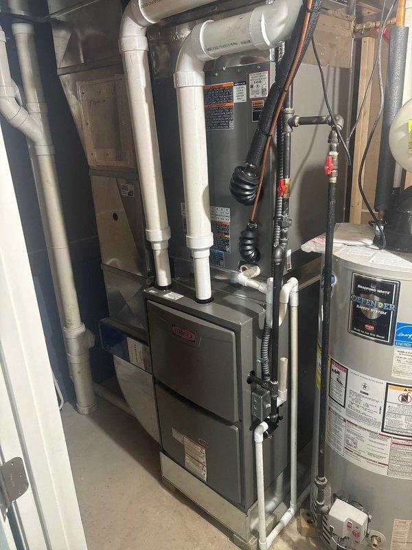 Completed furnace retrofit installation in residential mechanical room. Installed new dark gray cabinet-style furnace unit with white PVC venting system routed through ceiling, including multiple 90-degree elbows and straight sections for proper exhaust and intake air flow. Connected all necessary ductwork, copper piping with red-handle shut-off valves, and black flexible tubing to integrate with existing water heater and HVAC system components. Unit positioned on concrete floor with proper clearances maintained for service access and code compliance.