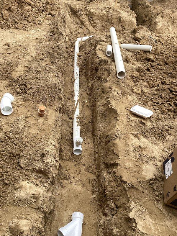 Completed rough-in plumbing installation for bathroom facilities at new construction site. Excavated trench using mini excavator in sandy soil conditions, installed white PVC drainage piping system including main lines, branch connections, and various fittings such as elbows and Y-connectors to accommodate multiple fixture locations. All pipes positioned and staged for final connection and testing prior to backfill.