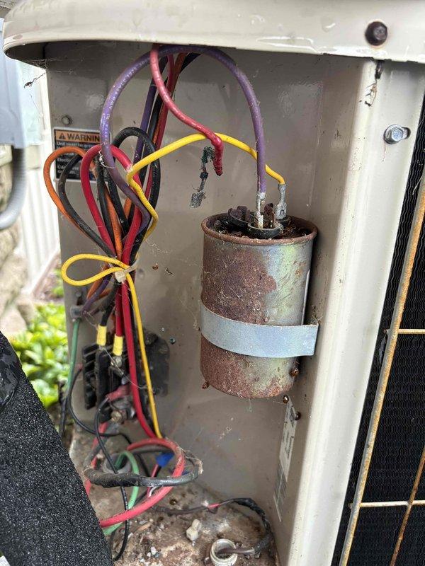 Completed cooling maintenance visit on residential air conditioning system. Upon inspection, discovered severely corroded and rusted capacitor within the electrical compartment, which posed a potential failure risk. The capacitor exhibited significant oxidation and deterioration from exposure to outdoor elements. Assessed all electrical connections and wiring, noting various colored conductors (red, yellow, purple, orange, and black) connected to the compromised component. Verified exterior condenser unit condition, which is positioned on a concrete pad with proper clearance and landscaping. Unit features standard white housing with top-mounted circular fan grille and metal mesh ventilation panels. Electrical disconnect box and conduit connections were examined for proper installation and safety compliance. Documented the corroded capacitor condition through photographs for service records and recommended immediate replacement to prevent system failure during peak cooling season.