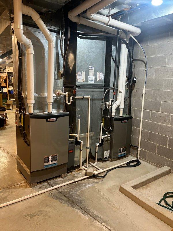 Completed full HVAC system replacement including installation of dual furnace units, central air handler, and two outdoor condensing units. Interior basement installation features organized PVC venting, copper refrigerant line sets, and integrated ductwork connections for multi-zone climate control. Exterior condensers positioned on prepared gravel pad with proper electrical disconnects, conduit routing, and refrigerant line connections secured to building exterior.