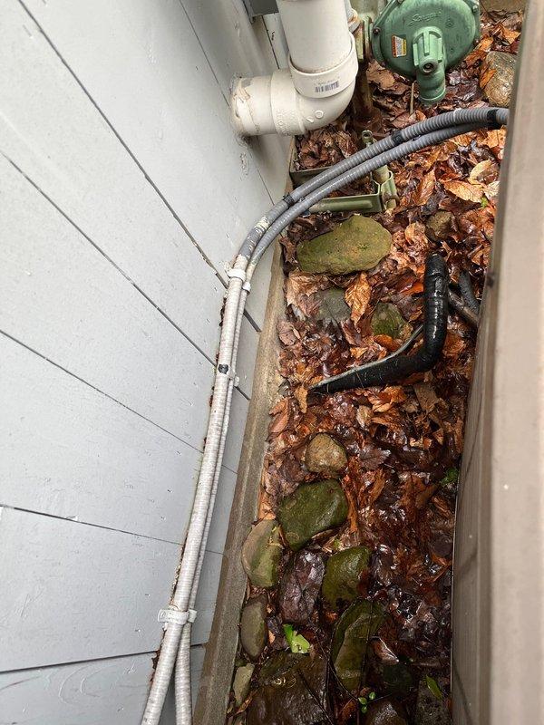 Completed AC replacement at residential property. Removed existing aged condenser unit and installed new system against exterior wall with light blue siding. New equipment positioned on existing rock pad base adjacent to utility area containing drainage piping and metering equipment. Verified proper clearances from building structure, plumbing components, and ground-level utilities. Connected refrigerant lines, electrical conduit, and condensate drainage to new unit. System tested and operational upon completion.