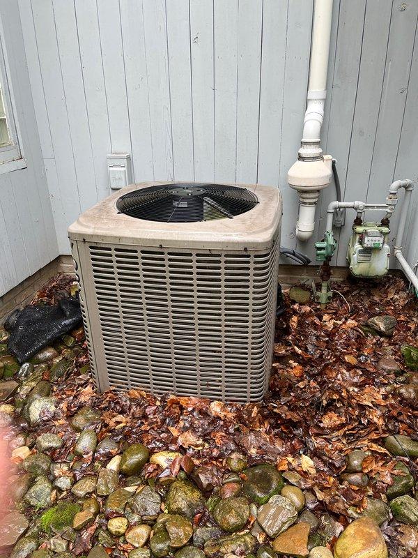 Completed AC replacement at residential property. Removed existing aged condenser unit and installed new system against exterior wall with light blue siding. New equipment positioned on existing rock pad base adjacent to utility area containing drainage piping and metering equipment. Verified proper clearances from building structure, plumbing components, and ground-level utilities. Connected refrigerant lines, electrical conduit, and condensate drainage to new unit. System tested and operational upon completion.
