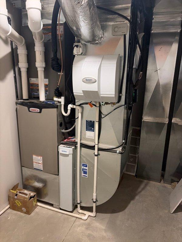 Completed new construction HVAC system installation including indoor furnace with integrated humidifier and outdoor condensing unit. The furnace was mounted in the basement utility area with proper venting and condensate drainage through white PVC piping, electrical connections secured with conduit, and flexible ductwork installed with foil tape sealing. The outdoor condensing unit was positioned on a concrete pad with gravel base adjacent to the stone foundation wall, ensuring proper clearances and airflow for optimal system performance.