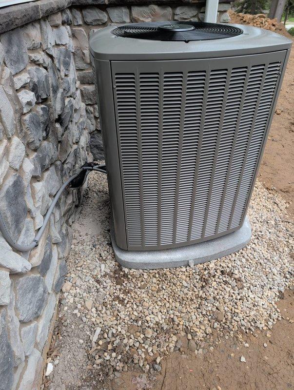 Completed new construction HVAC system installation including indoor furnace with integrated humidifier and outdoor condensing unit. The furnace was mounted in the basement utility area with proper venting and condensate drainage through white PVC piping, electrical connections secured with conduit, and flexible ductwork installed with foil tape sealing. The outdoor condensing unit was positioned on a concrete pad with gravel base adjacent to the stone foundation wall, ensuring proper clearances and airflow for optimal system performance.