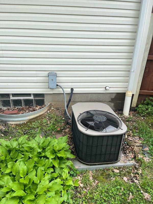 Completed AC retrofit installation at residential property. Exterior condenser unit installed on concrete pad against house exterior with proper electrical disconnect and conduit routing. Interior basement work included routing of refrigerant lines and associated piping through unfinished space to connect outdoor condensing unit to indoor air handling system.