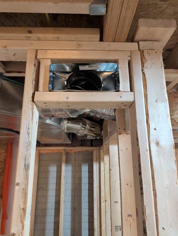 Completed new construction commercial HVAC installation including inline duct fan mounted within wooden chase assembly, metallic flex duct connections, and galvanized plenum box with proper sealing and insulation. All ductwork secured within framed opening with appropriate hangers and supports, system positioned for optimal airflow distribution throughout the space.