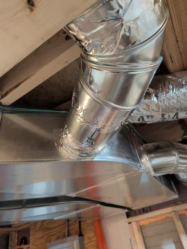Completed new construction commercial HVAC installation including inline duct fan mounted within wooden chase assembly, metallic flex duct connections, and galvanized plenum box with proper sealing and insulation. All ductwork secured within framed opening with appropriate hangers and supports, system positioned for optimal airflow distribution throughout the space.