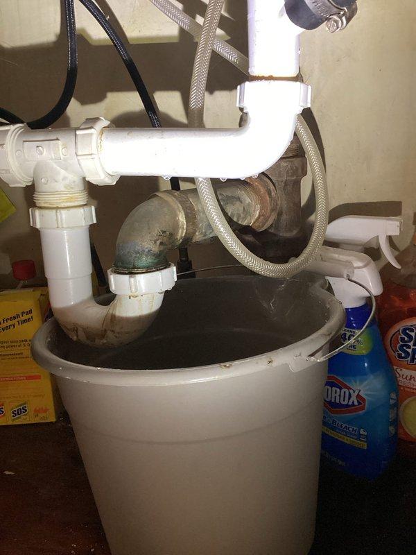Responded to service call for reported leak under kitchen sink. Upon arrival, inspected under-sink plumbing assembly and identified active leak at deteriorated metal elbow joint connection where it meets white PVC trap assembly. Observed water accumulation in bucket positioned beneath leak point, confirming ongoing drainage issue. 

Examined trap configuration and noted significant age-related deterioration on metal components, including visible oxidation and corrosion at threaded connections. Assessed condition of white PVC pipes and connections, observing discoloration and staining around joint areas indicating prolonged moisture exposure. Inspected braided flexible supply lines and confirmed they remained intact with no visible damage.

Conducted additional inspection of basement plumbing infrastructure to trace potential related issues. Examined exposed piping runs including PVC drain lines and supply pipes throughout basement ceiling joists. Verified no additional leak points or c
