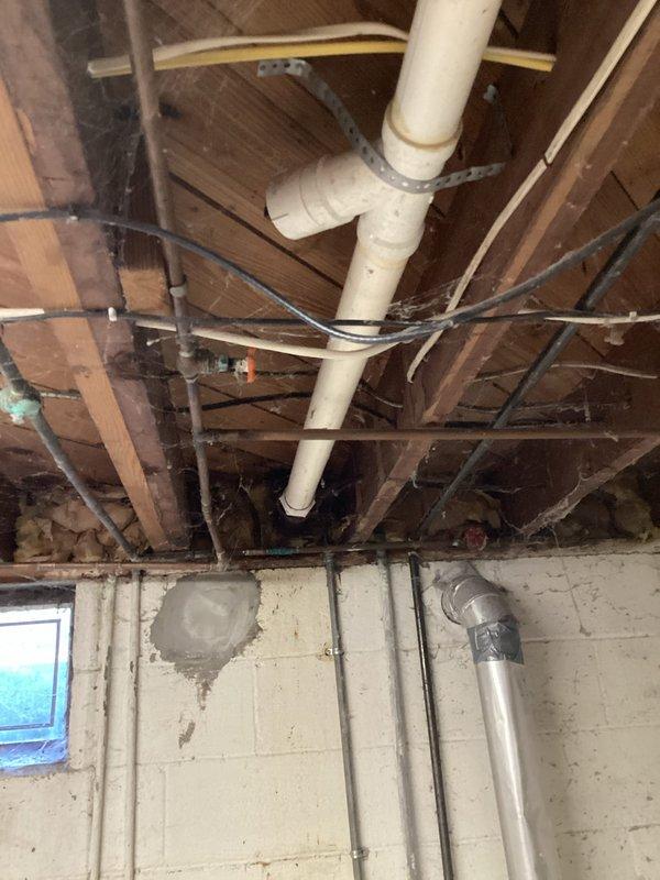 Responded to service call for reported leak under kitchen sink. Upon arrival, inspected under-sink plumbing assembly and identified active leak at deteriorated metal elbow joint connection where it meets white PVC trap assembly. Observed water accumulation in bucket positioned beneath leak point, confirming ongoing drainage issue. 

Examined trap configuration and noted significant age-related deterioration on metal components, including visible oxidation and corrosion at threaded connections. Assessed condition of white PVC pipes and connections, observing discoloration and staining around joint areas indicating prolonged moisture exposure. Inspected braided flexible supply lines and confirmed they remained intact with no visible damage.

Conducted additional inspection of basement plumbing infrastructure to trace potential related issues. Examined exposed piping runs including PVC drain lines and supply pipes throughout basement ceiling joists. Verified no additional leak points or c