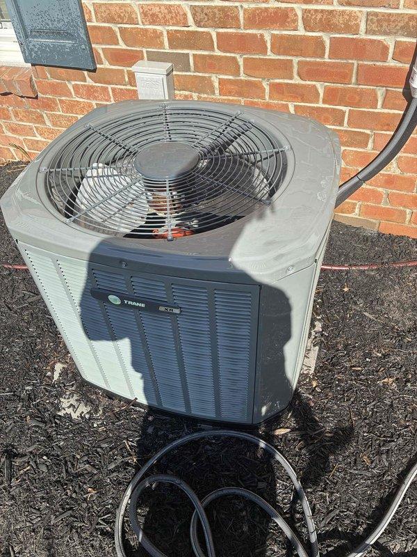 Completed comprehensive spring maintenance on Trane outdoor air conditioning condenser unit. Performed detailed inspection of all accessible components including capacitor, electrical connections, wiring harnesses, and refrigerant lines. Verified proper operation of condenser fan assembly and protective guard, checked electrical connections with red wire nuts for security, and inspected the condition of the cylindrical capacitor with all terminal connections. Examined exterior cabinet for weathering and structural integrity, confirmed refrigerant lines and electrical conduit properly secured along building exterior, and verified unit placement on pad with adequate clearance from brick wall. All components found to be in functional operating condition with normal seasonal wear patterns observed on exterior surfaces.