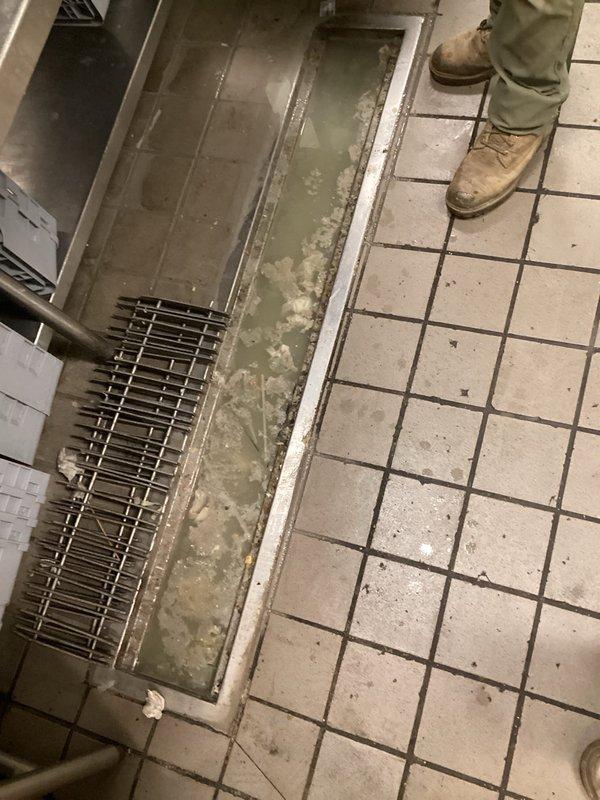 # Commercial Kitchen Floor Drain Cleaning and Maintenance

Responded to a clogged commercial kitchen floor drain system with standing water and visible debris accumulation. Upon arrival, assessed a rectangular drainage trough running through the center of white tile flooring, which contained discolored standing water with greenish-brown contamination and significant debris buildup.

Removed metal grating covering the drainage channel to access the obstruction. Cleared accumulated food waste, paper debris, and organic matter from both the grate surface and drain channel. Extracted blockage materials causing water backup in the trough system. Flushed the drain line to restore proper water flow and drainage capacity.

Cleaned the metal grate components and reinstalled them securely. Verified drainage system was flowing freely with no standing water remaining. Recommended regular maintenance schedule to prevent future buildup in this high-traffic commercial kitchen environment.