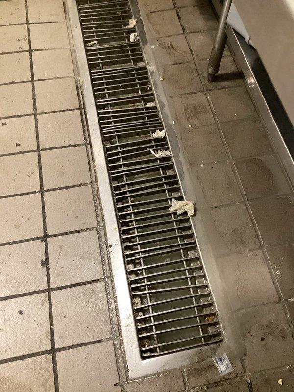 # Commercial Kitchen Floor Drain Cleaning and Maintenance

Responded to a clogged commercial kitchen floor drain system with standing water and visible debris accumulation. Upon arrival, assessed a rectangular drainage trough running through the center of white tile flooring, which contained discolored standing water with greenish-brown contamination and significant debris buildup.

Removed metal grating covering the drainage channel to access the obstruction. Cleared accumulated food waste, paper debris, and organic matter from both the grate surface and drain channel. Extracted blockage materials causing water backup in the trough system. Flushed the drain line to restore proper water flow and drainage capacity.

Cleaned the metal grate components and reinstalled them securely. Verified drainage system was flowing freely with no standing water remaining. Recommended regular maintenance schedule to prevent future buildup in this high-traffic commercial kitchen environment.