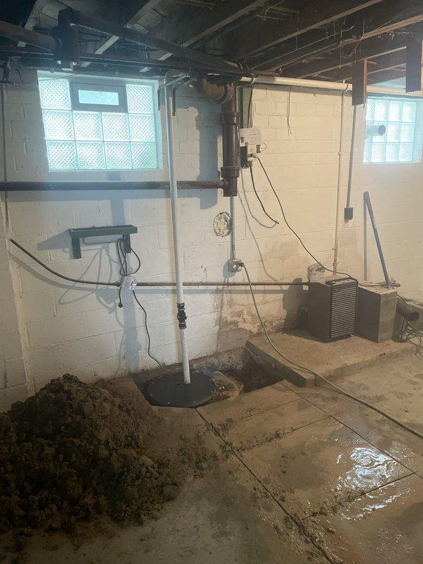 Performed concrete floor excavation in basement to provide access for new sewer line installation. Created rectangular opening in concrete foundation, exposing existing underground piping system that required replacement. Removed soil and debris to establish proper depth and clearance for new sewer line routing into the residence, with work area prepared for subsequent pipe installation and connection to main system.