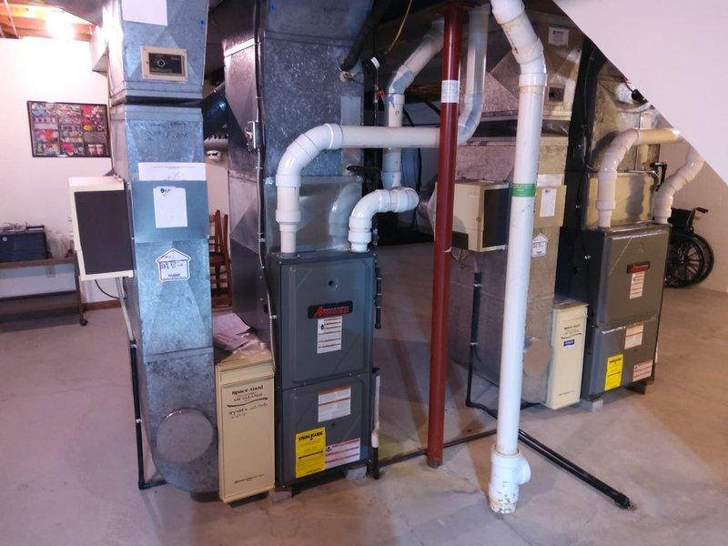 Completed comprehensive HVAC estimate for AC retrofit installation in residential basement mechanical room. Assessed existing furnace and air handling equipment, evaluated ductwork configuration including gray return air ducts and overhead galvanized steel components, and documented current PVC condensate drainage system with white piping running to floor drain. Photographed existing multi-unit HVAC setup, noting wall-mounted control panel, thermostat placement, and exposed ceiling joist structure to determine optimal retrofit approach for new air conditioning system integration with current heating infrastructure.