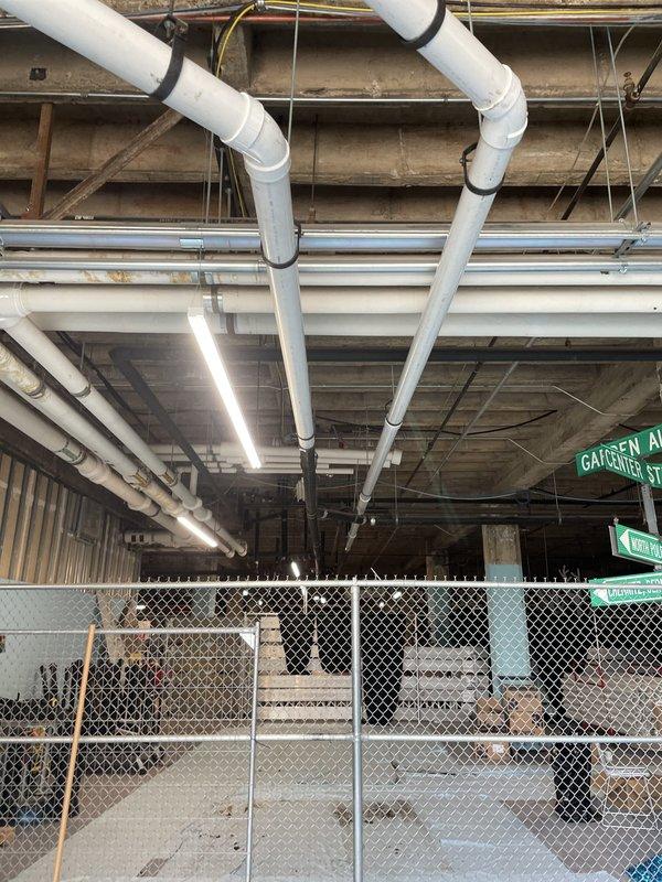 Completed installation of large-diameter white PVC drainage piping system in commercial space beneath elevated structure, routing pipes across exposed concrete ceiling with proper securing brackets and support straps. Installed parallel gray metallic piping runs and coordinated routing with existing fluorescent lighting fixtures and structural elements to ensure proper clearance and code compliance.