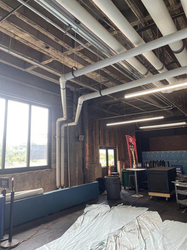 Completed installation of large-diameter white PVC drainage piping system in commercial space beneath elevated structure, routing pipes across exposed concrete ceiling with proper securing brackets and support straps. Installed parallel gray metallic piping runs and coordinated routing with existing fluorescent lighting fixtures and structural elements to ensure proper clearance and code compliance.