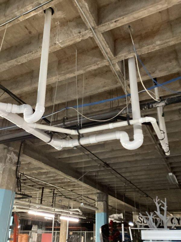 Completed installation of white PVC drainage piping system in commercial building ceiling space, including multiple horizontal runs, vertical drops, and U-shaped configurations to establish proper drainage flow. Installed support wiring and hangers to secure piping to exposed concrete ceiling structure, ensuring all connections meet code requirements for commercial plumbing applications.
