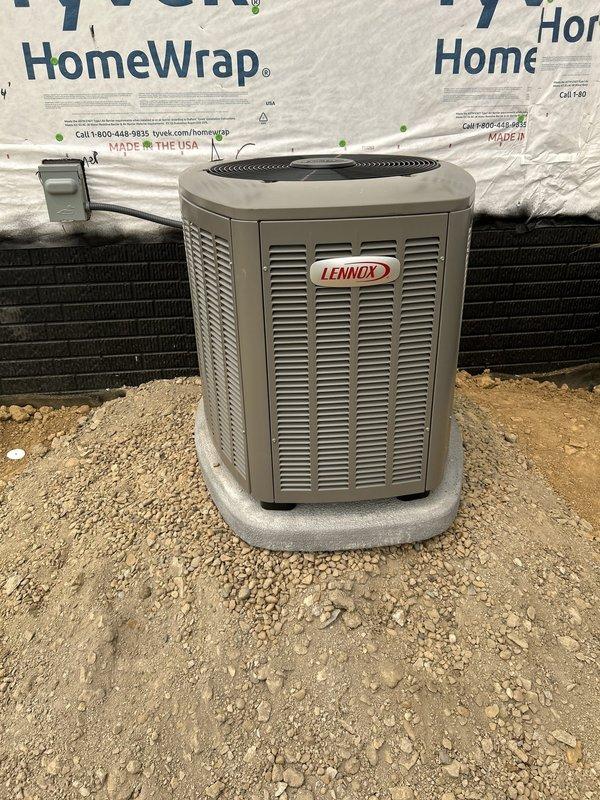 # HVAC New Construction Install

Completed installation of dual Lennox outdoor condenser units on new construction residential property. Positioned two condenser units side by side on gravel base with proper composite equipment pads for stability and drainage. Units include one black condenser and one beige/tan Lennox-branded unit, both mounted against exterior wall with Tyvek house wrap. Installed insulated refrigerant line sets with proper bending radius from top of each unit. Mounted electrical disconnect box to wall for code compliance and service accessibility. Equipment properly leveled on crushed stone base to ensure optimal performance and drainage. All refrigerant lines routed and secured according to manufacturer specifications.