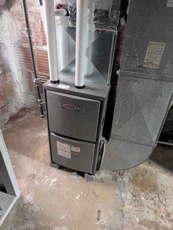 Completed furnace retrofit installation in residential basement utility space. Installed new gray Lennox furnace unit with dual white PVC intake and exhaust venting system running vertically from top of unit. Connected copper refrigerant lines, integrated with existing green air handler cabinet, and installed galvanized metal ductwork with proper sealing and support. All manufacturer specifications verified, digital control panel programmed and operational, system tested and confirmed functioning within normal parameters.