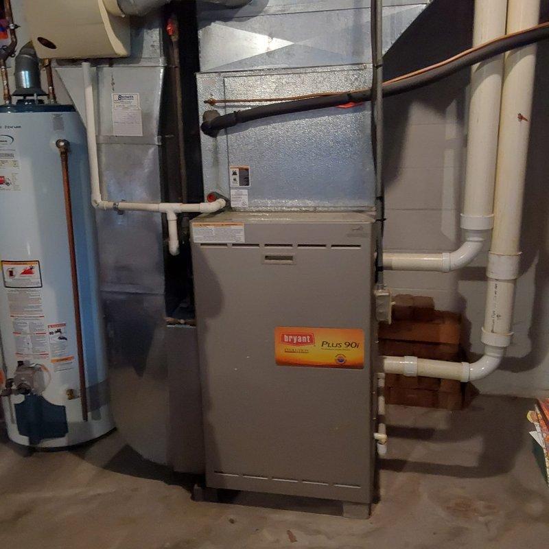 Completed comprehensive HVAC system evaluation for air conditioning retrofit at residential property. Assessed existing outdoor condenser unit positioned against exterior brick wall, noting vegetation growth around equipment that required clearance consideration for proper airflow and maintenance access. Documented indoor basement mechanical room containing Bryant Plus 90i furnace and existing water heater with associated copper piping, PVC venting system, and ductwork infrastructure. Evaluated compatibility of current furnace system with proposed air conditioning retrofit, inspected electrical panel accessibility, and confirmed adequate space for refrigerant line routing between indoor and outdoor components. Provided detailed photographic documentation of both outdoor condenser location and indoor mechanical room configuration to support equipment replacement specifications and installation planning.