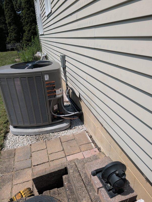 Completed full HVAC system retrofit including outdoor air conditioning condenser unit and indoor vertical air handler installation. The outdoor condenser was positioned on a gravel pad against the building exterior with proper electrical conduit and refrigerant line routing through the siding, while the indoor air handler was installed in the basement mechanical room with integrated PVC condensate drainage system and pump. All components were properly connected, tested, and verified operational per manufacturer specifications.