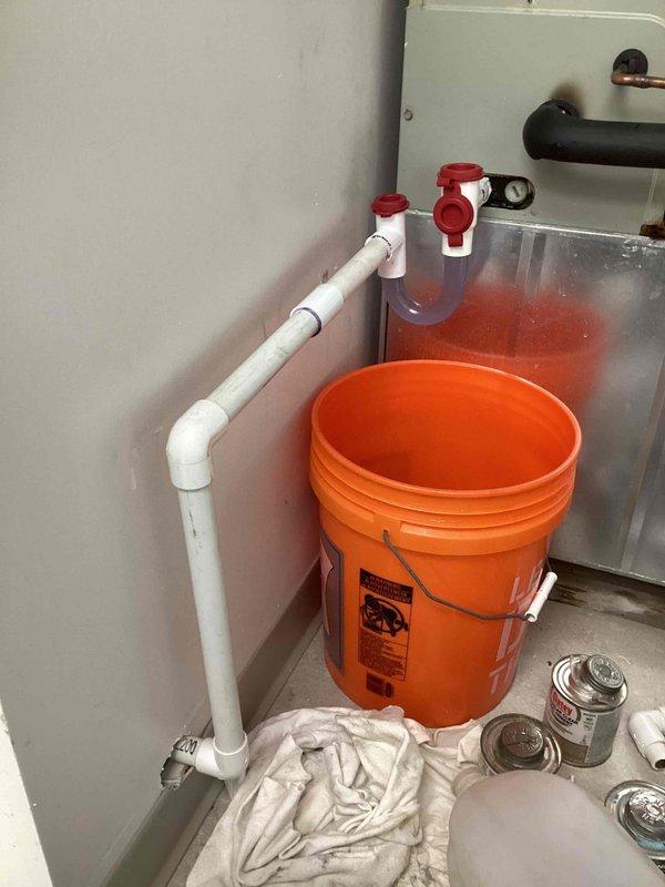 # Commercial A/C Service - Condensate Drainage Emergency

Responded to emergency service call for active water discharge from commercial air conditioning system. Customer reported recurring issue identical to problem experienced two years prior, with water gushing from unit.

Upon arrival, found significant condensate overflow condition requiring immediate attention. Inspected existing PVC condensate drainage system running along wall with multiple elbow joints and fittings. Examined air handler unit and associated HVAC equipment installation in mechanical room.

Identified condensate drain line blockage causing water backup and overflow. Placed collection bucket to contain active water discharge and prevent further property damage. Assessed drain line routing, connections, and termination points throughout system.

Cleared obstruction from condensate drain line using appropriate tools and techniques. Verified proper drainage flow and tested system operation under load. Inspected drain