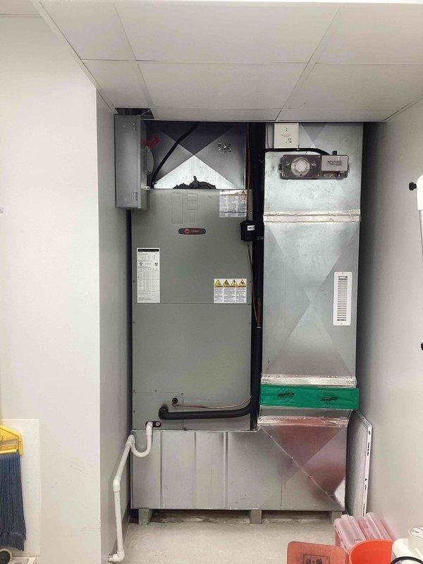 # Commercial A/C Service - Condensate Drainage Emergency

Responded to emergency service call for active water discharge from commercial air conditioning system. Customer reported recurring issue identical to problem experienced two years prior, with water gushing from unit.

Upon arrival, found significant condensate overflow condition requiring immediate attention. Inspected existing PVC condensate drainage system running along wall with multiple elbow joints and fittings. Examined air handler unit and associated HVAC equipment installation in mechanical room.

Identified condensate drain line blockage causing water backup and overflow. Placed collection bucket to contain active water discharge and prevent further property damage. Assessed drain line routing, connections, and termination points throughout system.

Cleared obstruction from condensate drain line using appropriate tools and techniques. Verified proper drainage flow and tested system operation under load. Inspected drain